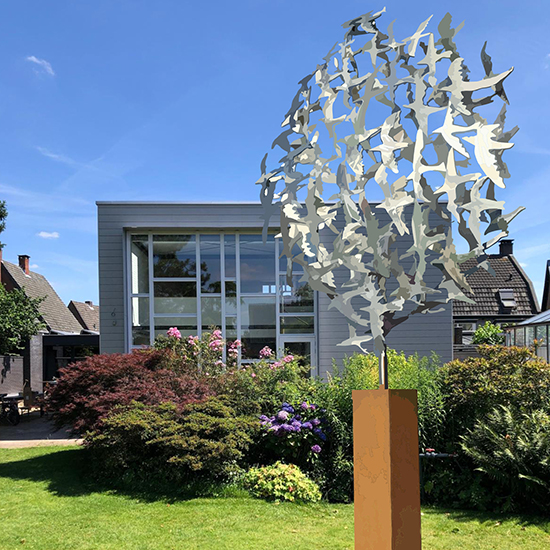 Rond metalen beeld met uitsparingen van vogelfiguren op een cortenstalen sokkel in een tuin.