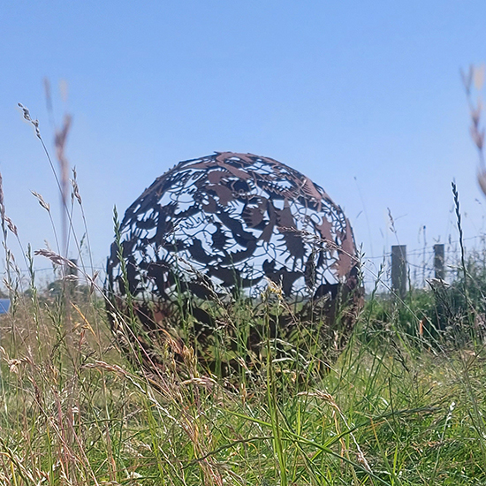Balvormig cortenstalen beeld met uitsparingen van vogelvormen, liggend in het gras.