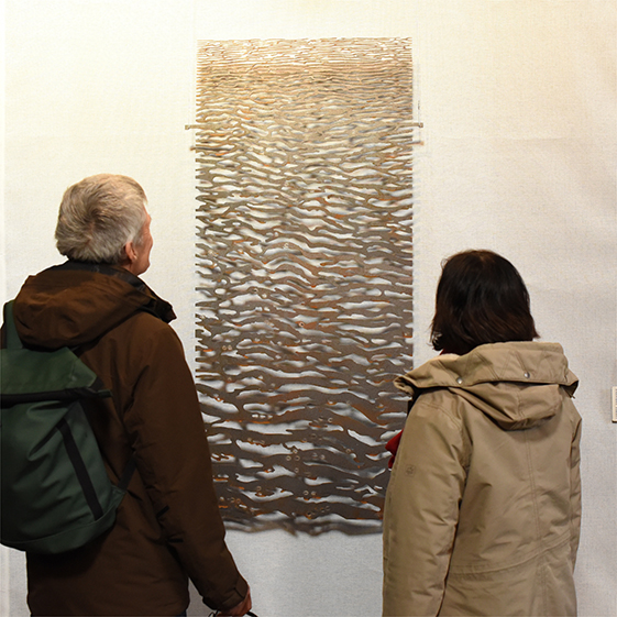 Twee mensen bekijken een hangend beeld van staal met golvende uitsparingen.