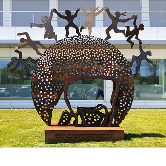 Rond cortenstalen beeld met gaten en een uitgesneden koe, met dansende mensfiguren bovenop.