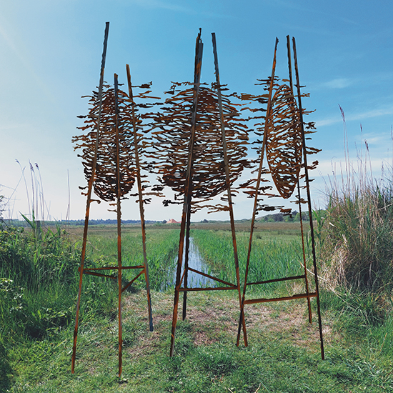 Drie op cortenstalen palen staande ronde beelden van cortenstaal met uitsparingen geïnspireerd op zee, vloed en nesten, geplaatst in een weiland.