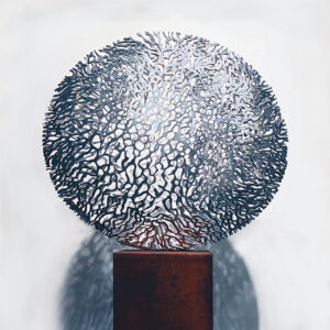 Rondvormige metalen beeld met organische uitsparingen, geplaatst op een sokkel.