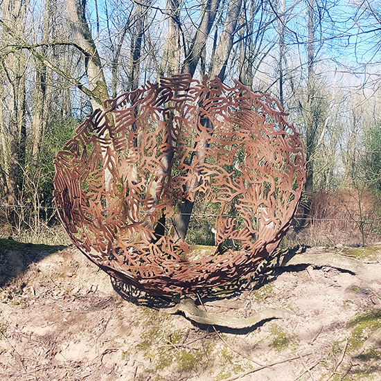 Schaalvormig cortenstalen beeld met organische, nautisch geïnspireerde uitsparingen, liggend op een heuvel voor een rij bomen.